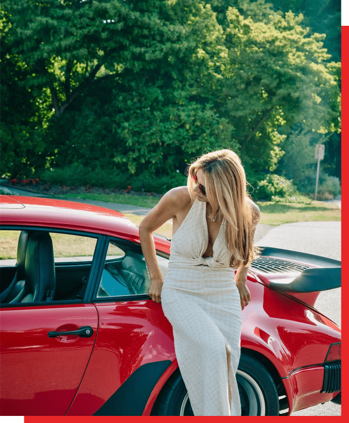 A woman in white dress leaning on the hood of a red car.